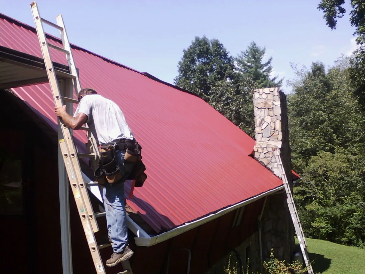 Roofer on a ladder beside a red metal roof and stone chimney — Soffit & Fascia Repair by Roofing Brothers in Orangeville